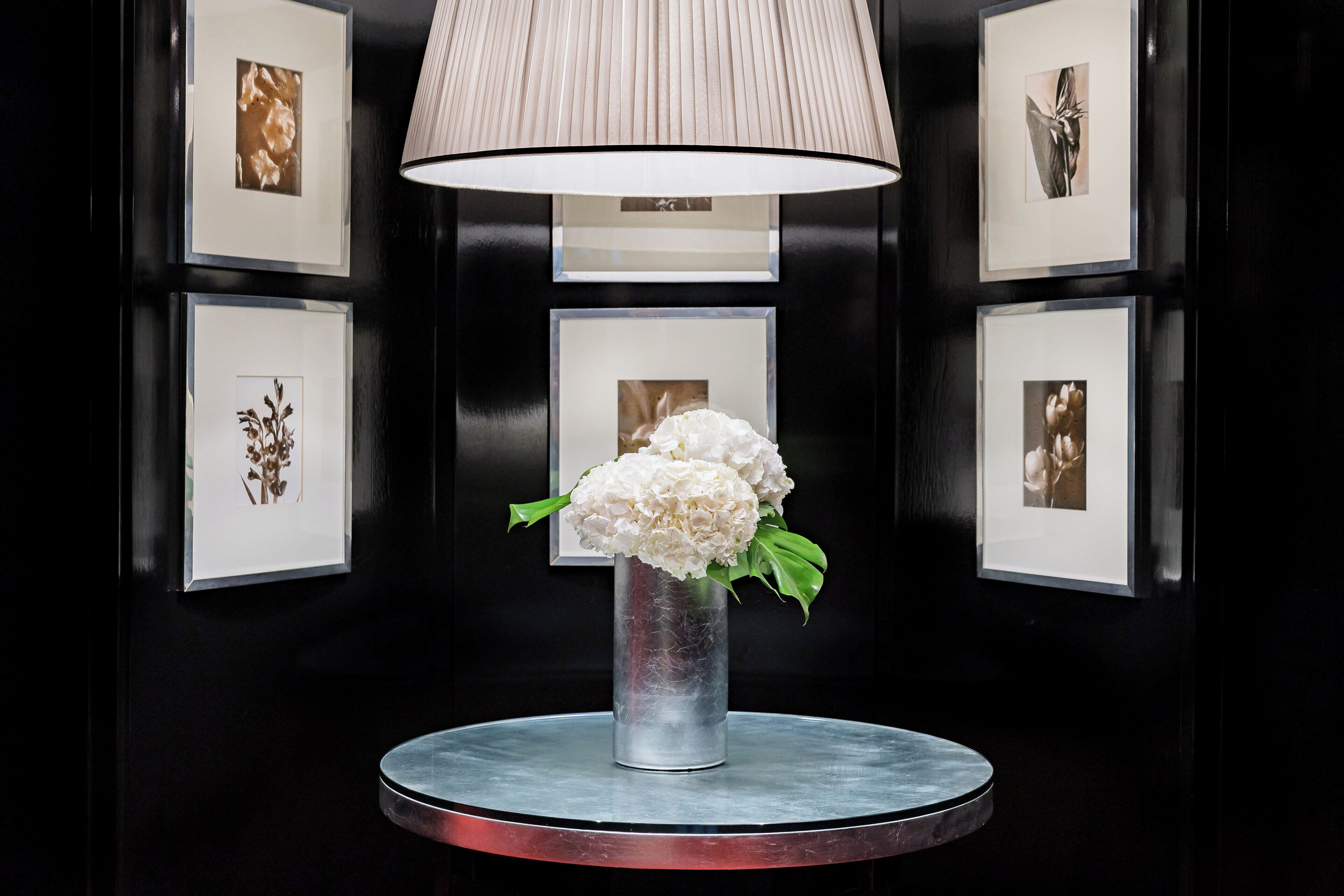 A black-framed wall with six framed photographs and a vase with white flowers on a round table.