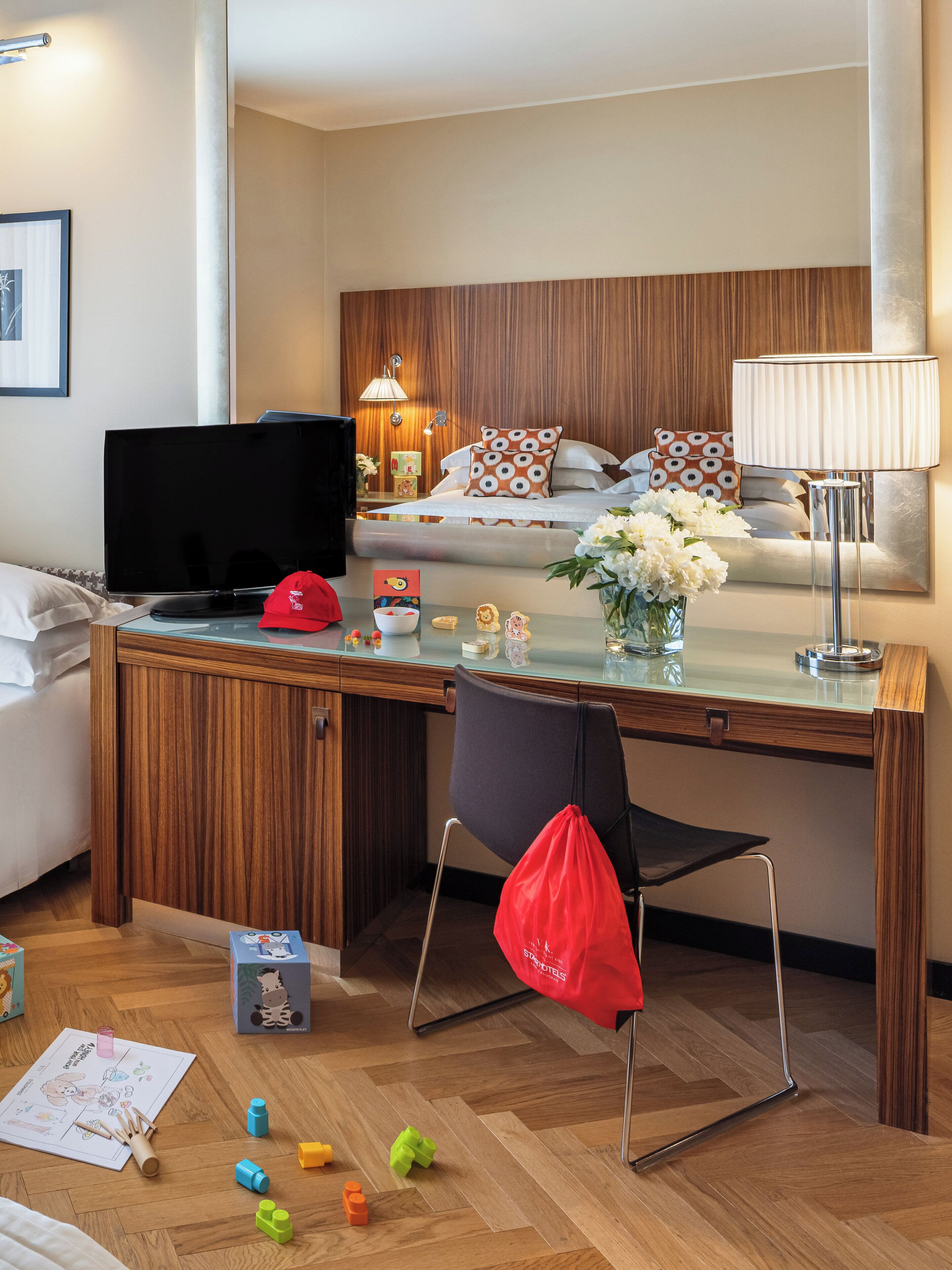 Une chambre d’hôtel avec un bureau en bois, une télévision, un lit avec des coussins à motifs, une lampe de chevet et un vase rempli de fleurs.