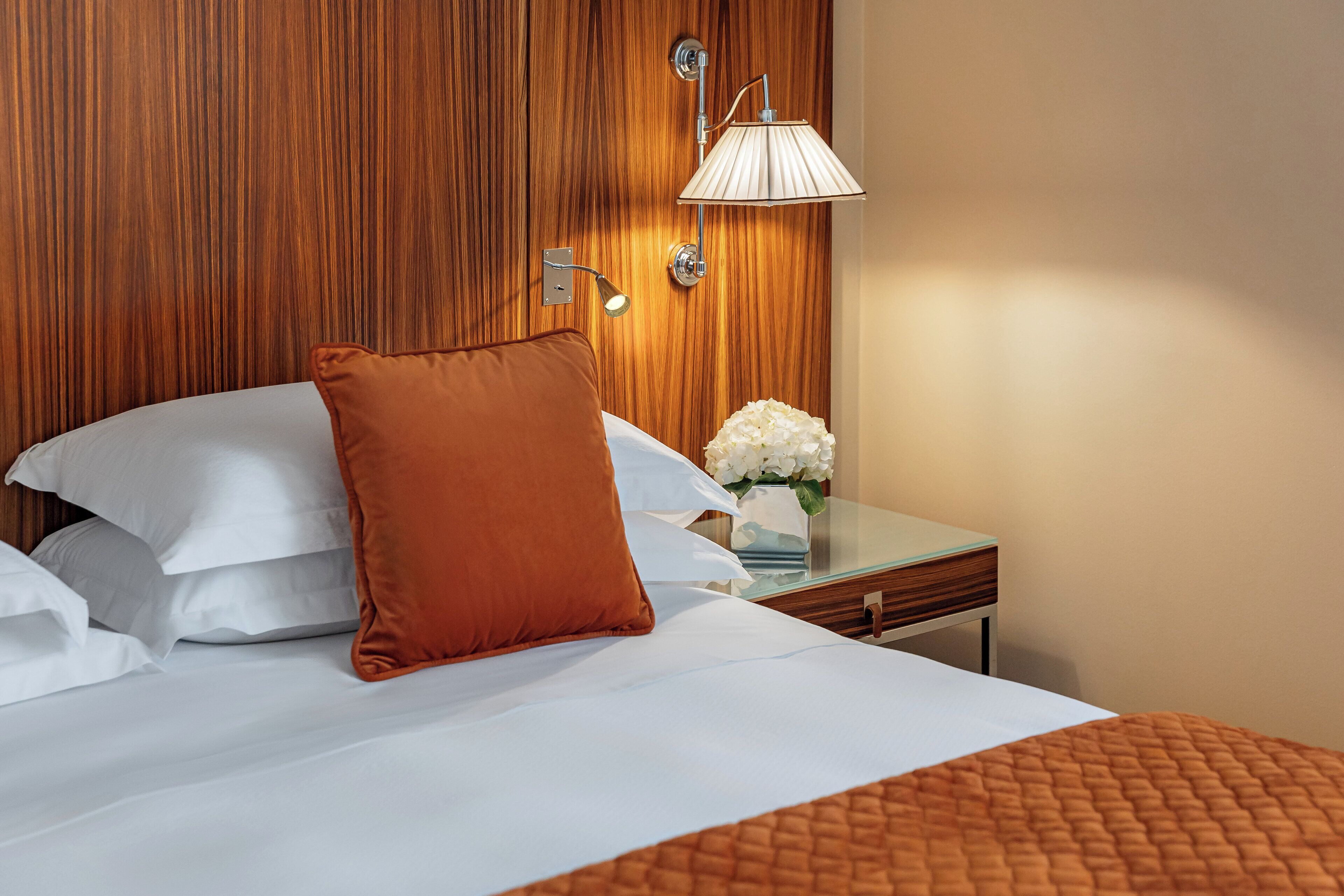 A neatly made bed with white and brown pillows, a glass side table with a vase of white flowers, and a wooden headboard.