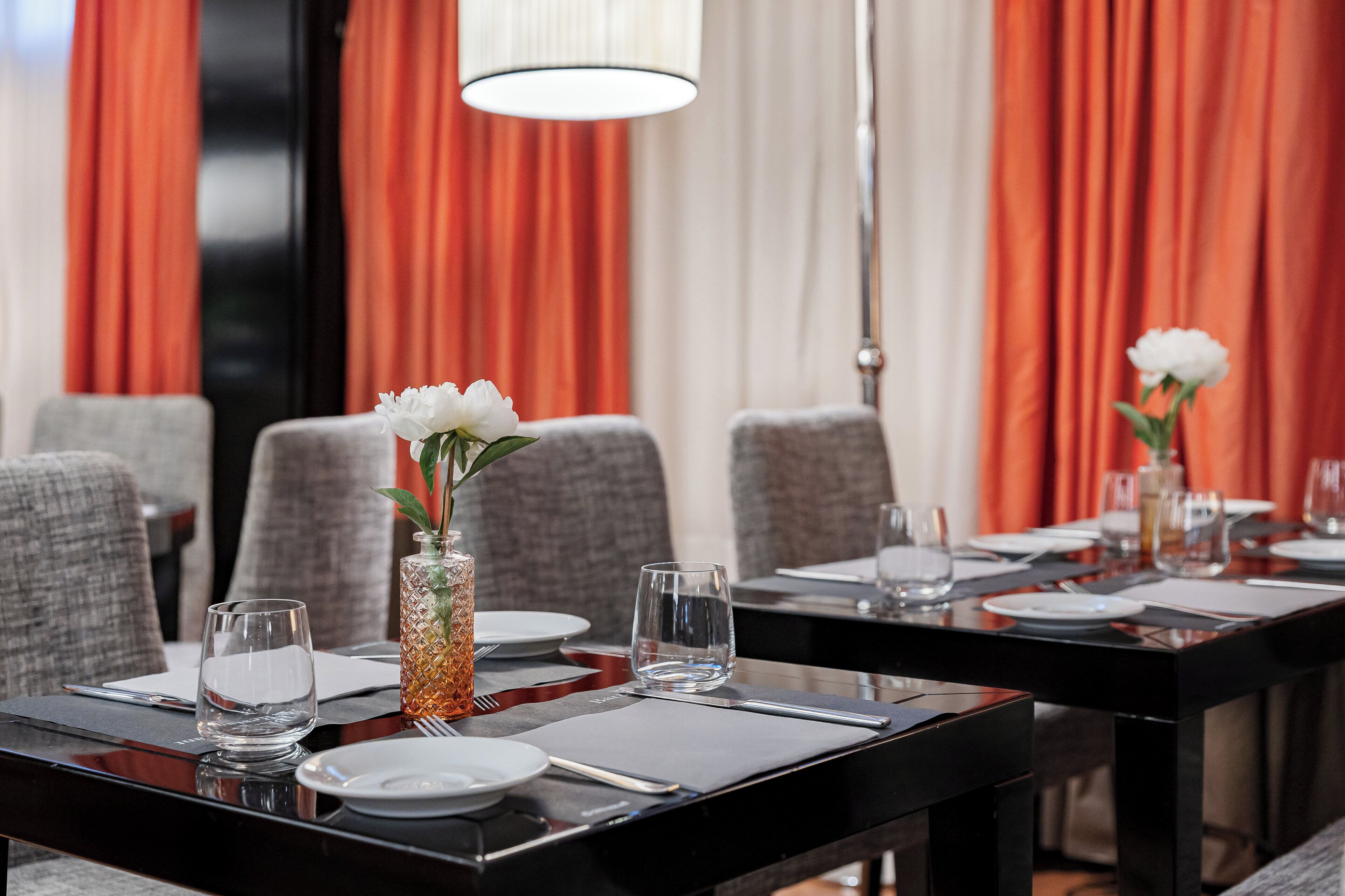 Une table à manger dressée avec des assiettes, des verres et des couverts, entourée de chaises, dans une pièce aux rideaux rouges et ornée d’un luminaire suspendu.