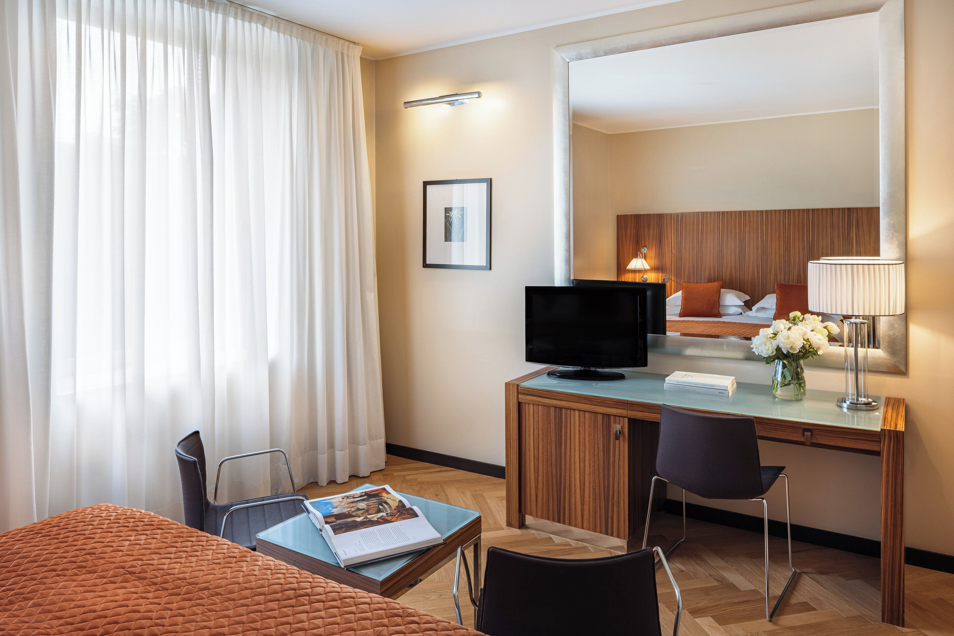 Et hotelværelse med seng, skrivebord med tv, stol, lampe og et vindue med gardiner.