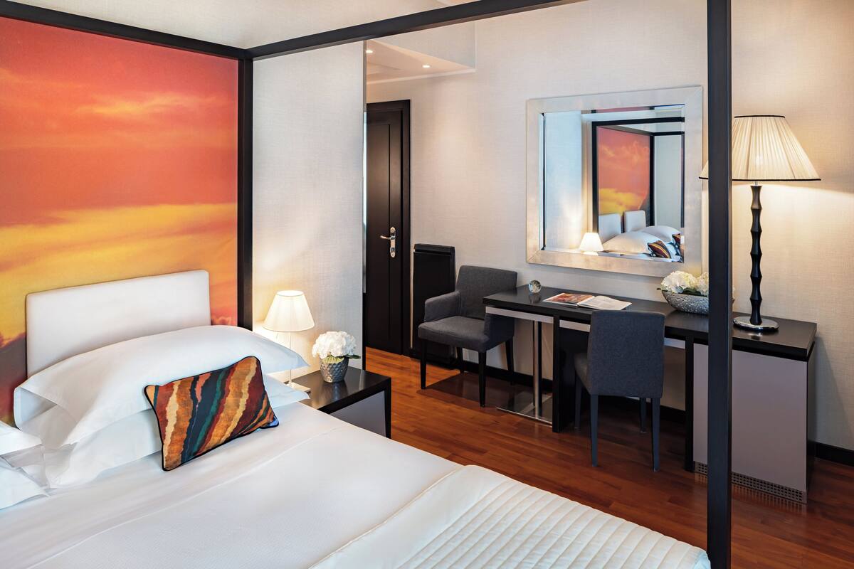 Hotelkamer met een bed, bureau, stoel, bureaulamp, spiegel en een muur met een zonsondergangschildering.