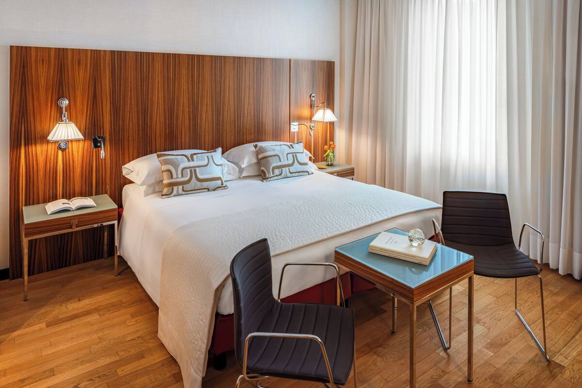 Een hotelkamer met een houten hoofdeinde, een groot bed met wit beddengoed, twee bedlampen, een klein tafeltje met een boek, en twee stoelen.