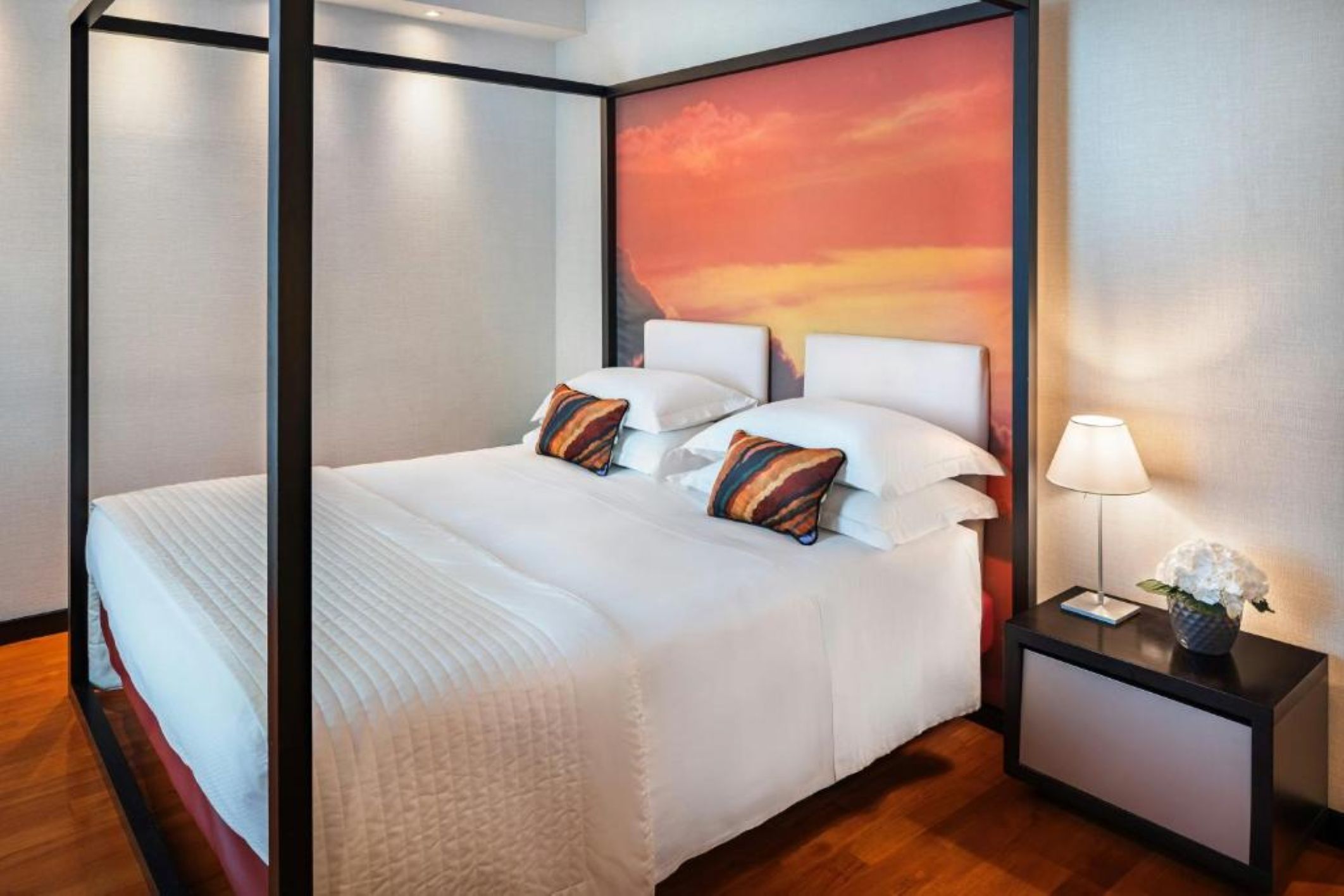 A four-poster bed with white bedding, a nightstand with a lamp, and a painting of a sunset on the wall.
