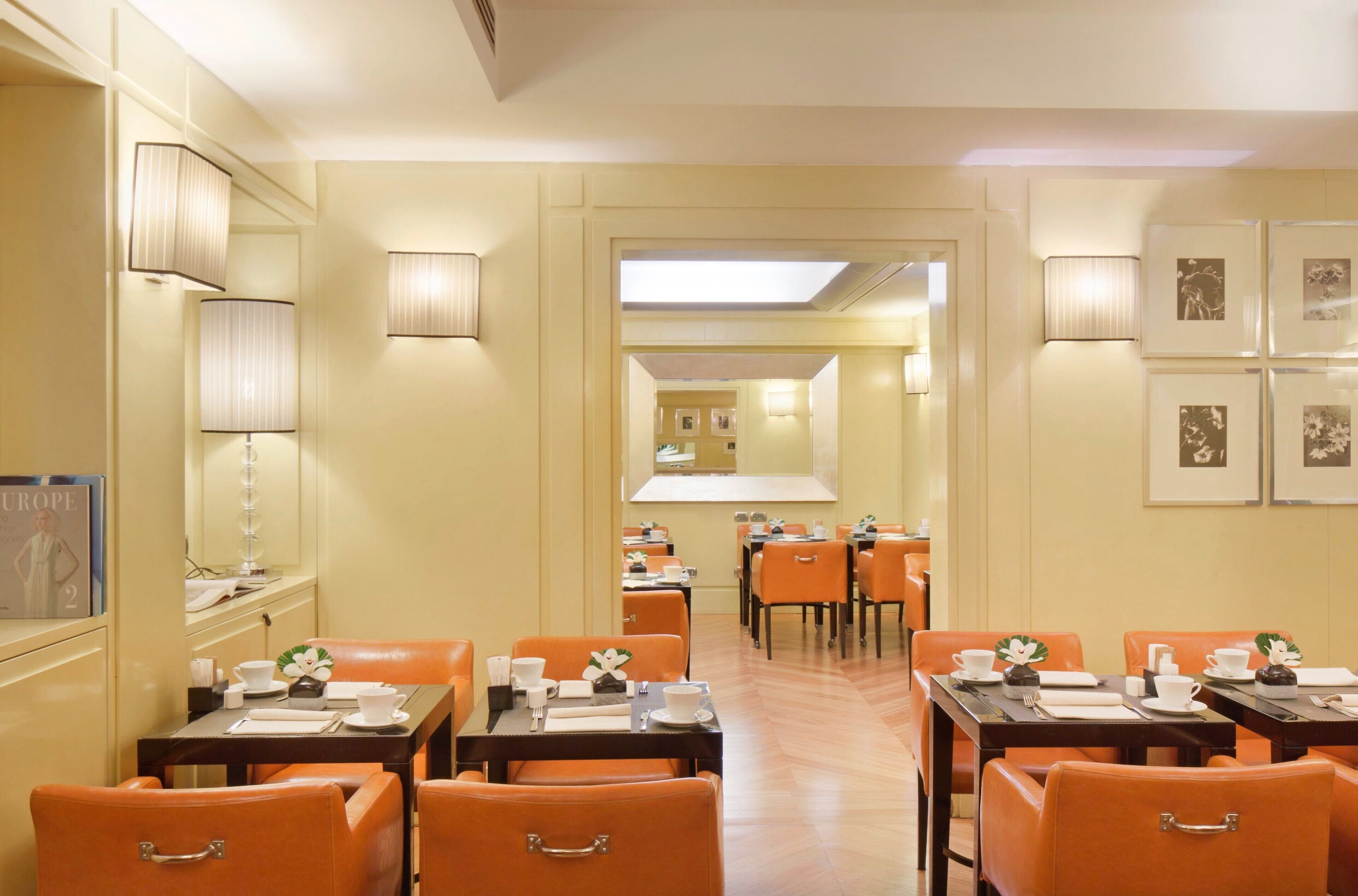 Uma área de refeições de hotel com cadeiras laranja, mesas montadas com louça branca e quadros nas paredes.