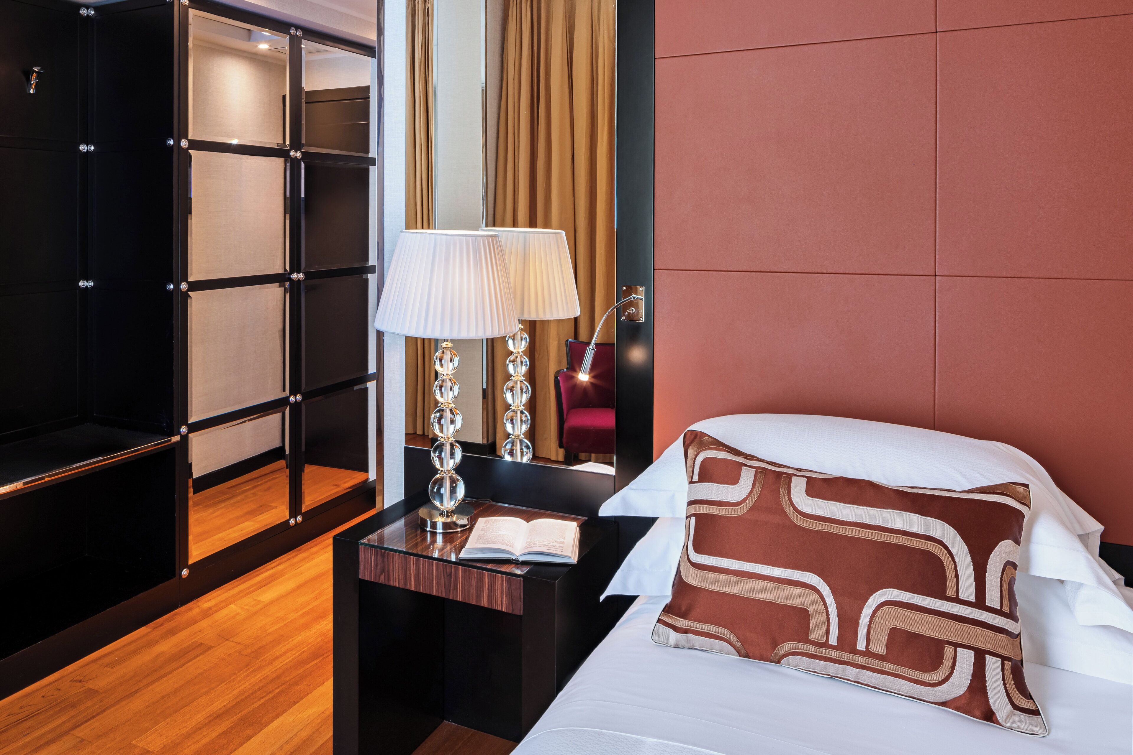 Quarto de hotel com cama, mesa de cabeceira, abajur, um livro e guarda-roupa.