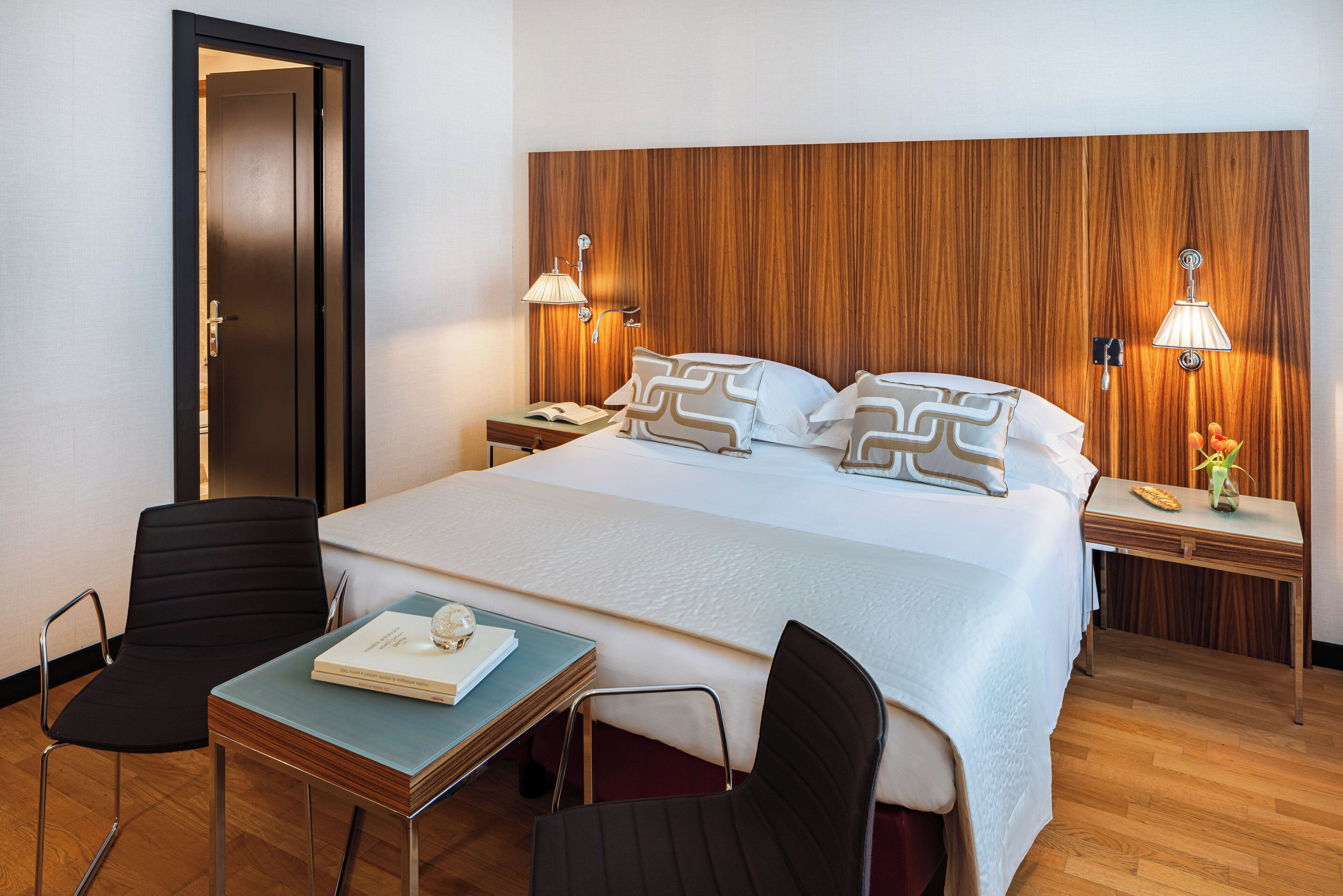 A hotel room with a large bed, two bedside tables, a nightstand, a desk with a chair, and a door.
