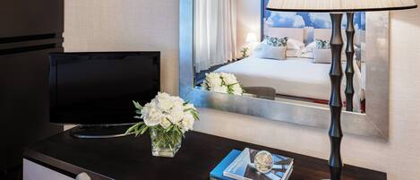 Una habitación de hotel con una cama, televisión, escritorio con lámpara, un florero con flores y un libro.