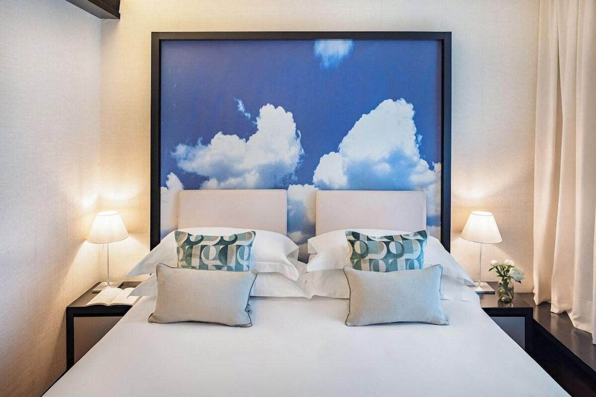 A hotel room with a large bed, two bedside tables, and a wall-mounted artwork of clouds.
