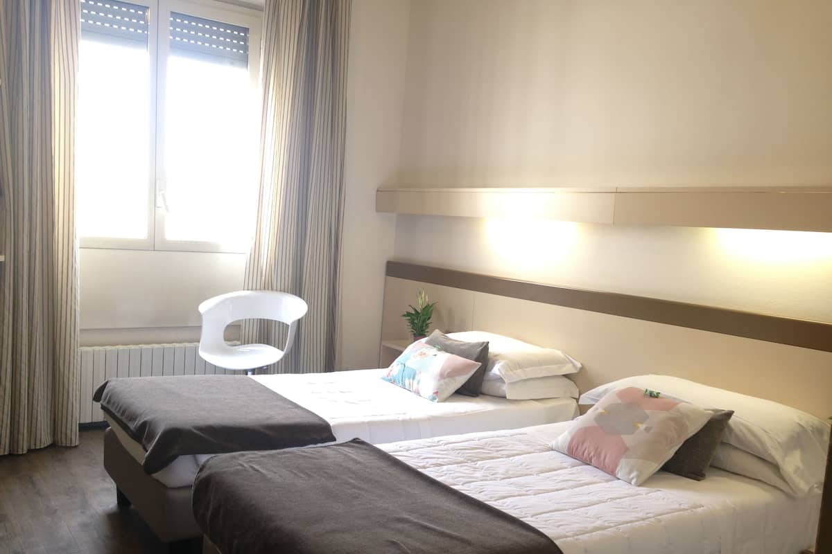 A hotel room with two beds, a bedside table, a chair, and a window with curtains.