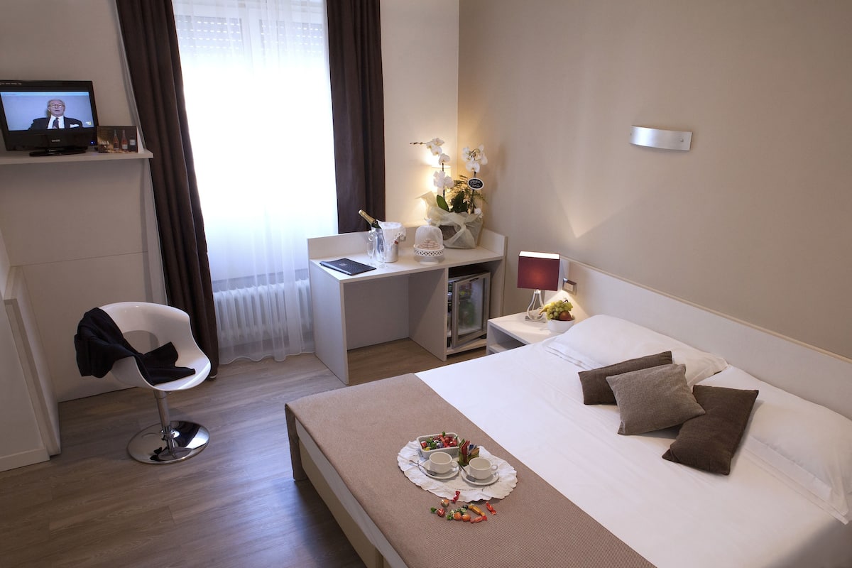A hotel room with a bed, a desk, a chair, a TV, and a bedside table with a lamp and a vase of flowers.