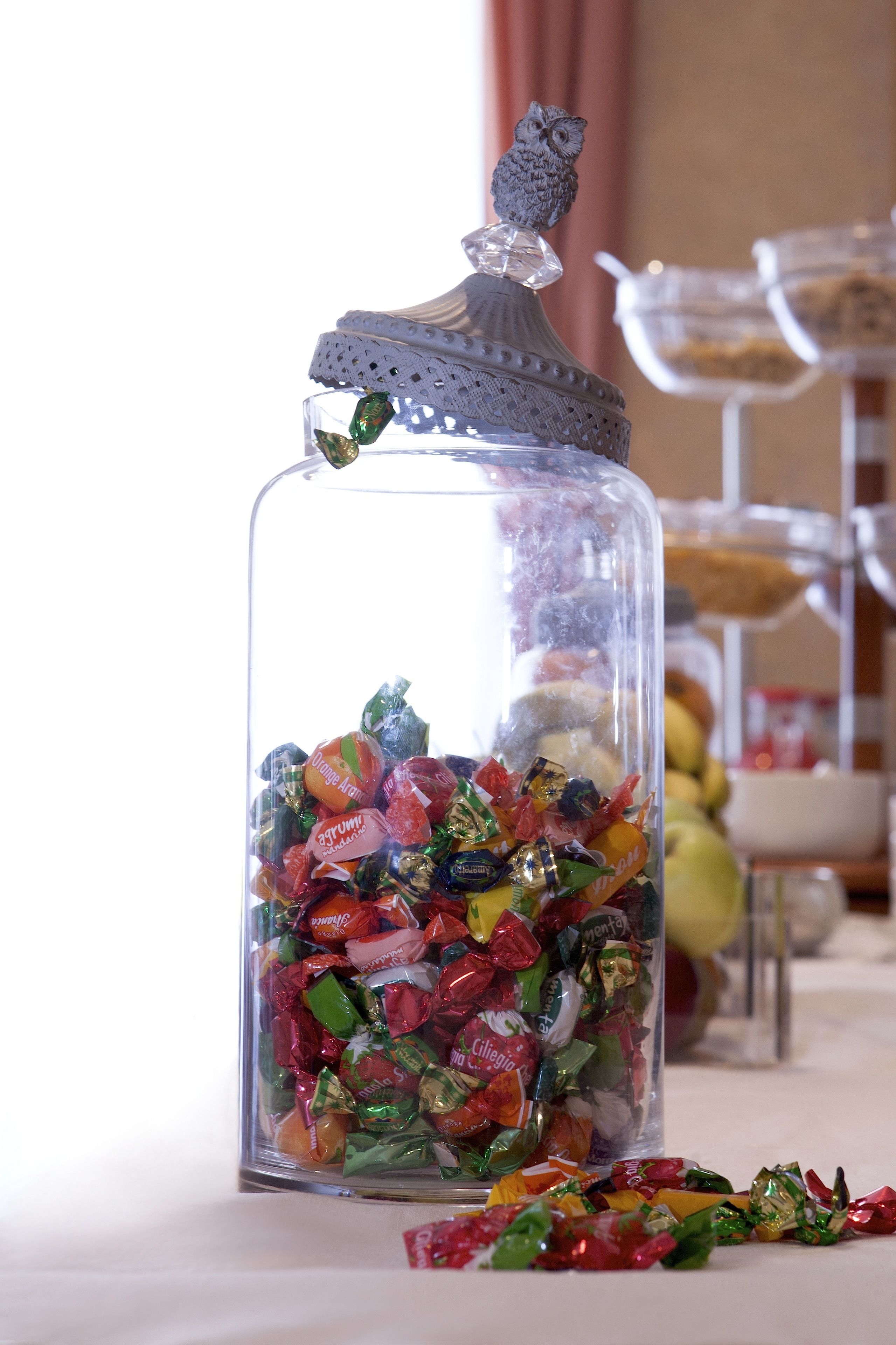A glass jar filled with assorted candies, placed on a table with a decorative owl on top.