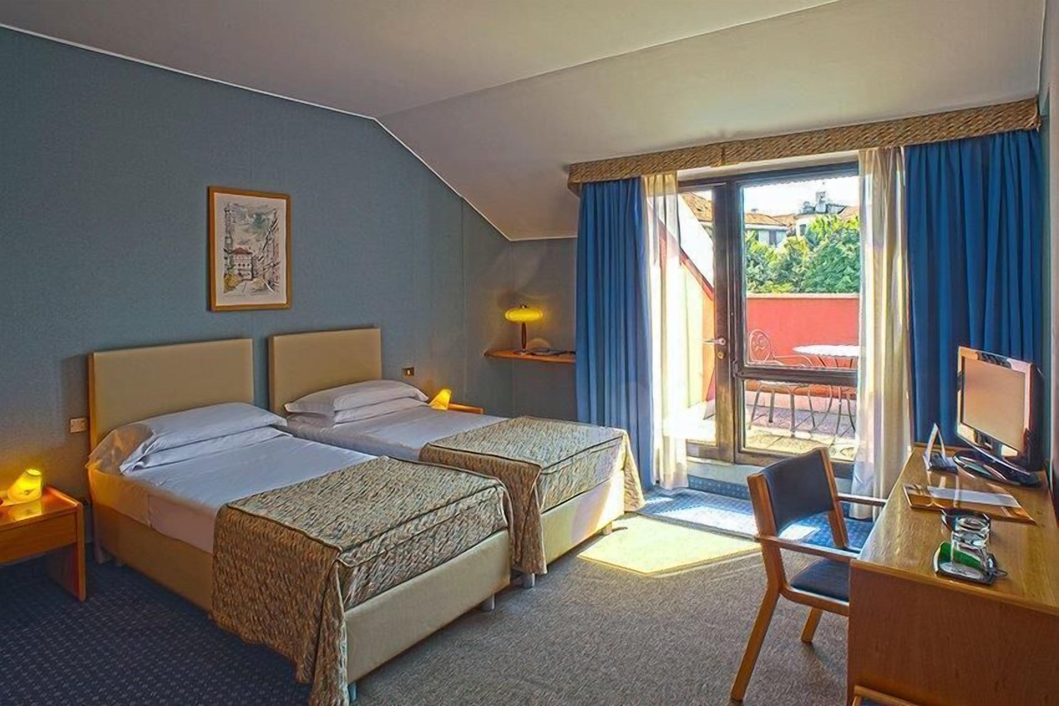 A hotel room with two beds, a desk with a computer, a chair, a small table with a lamp, and a window with curtains.