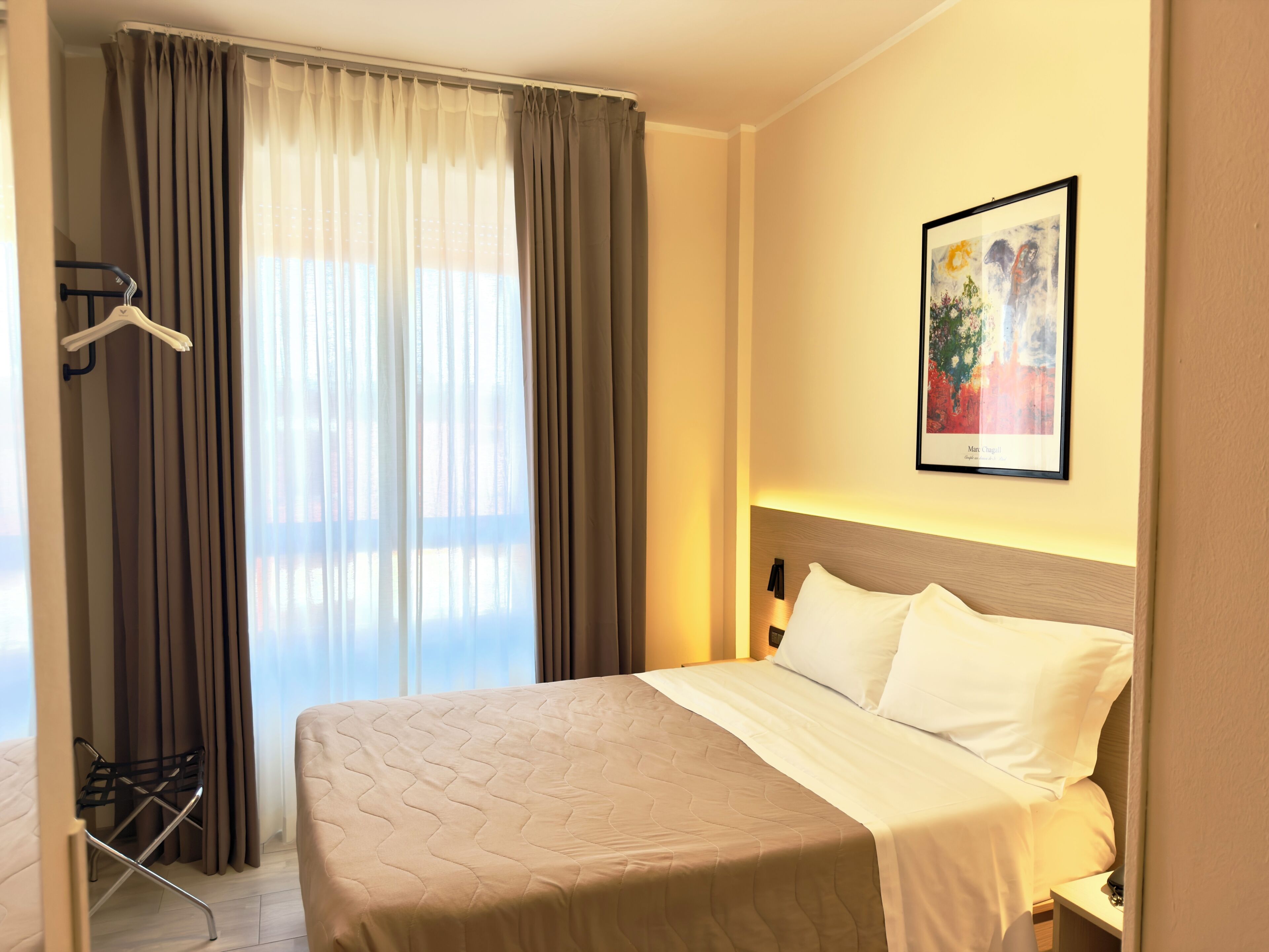 Quarto de hotel com cama, janela com cortinas, quadro na parede e uma pequena mesa.