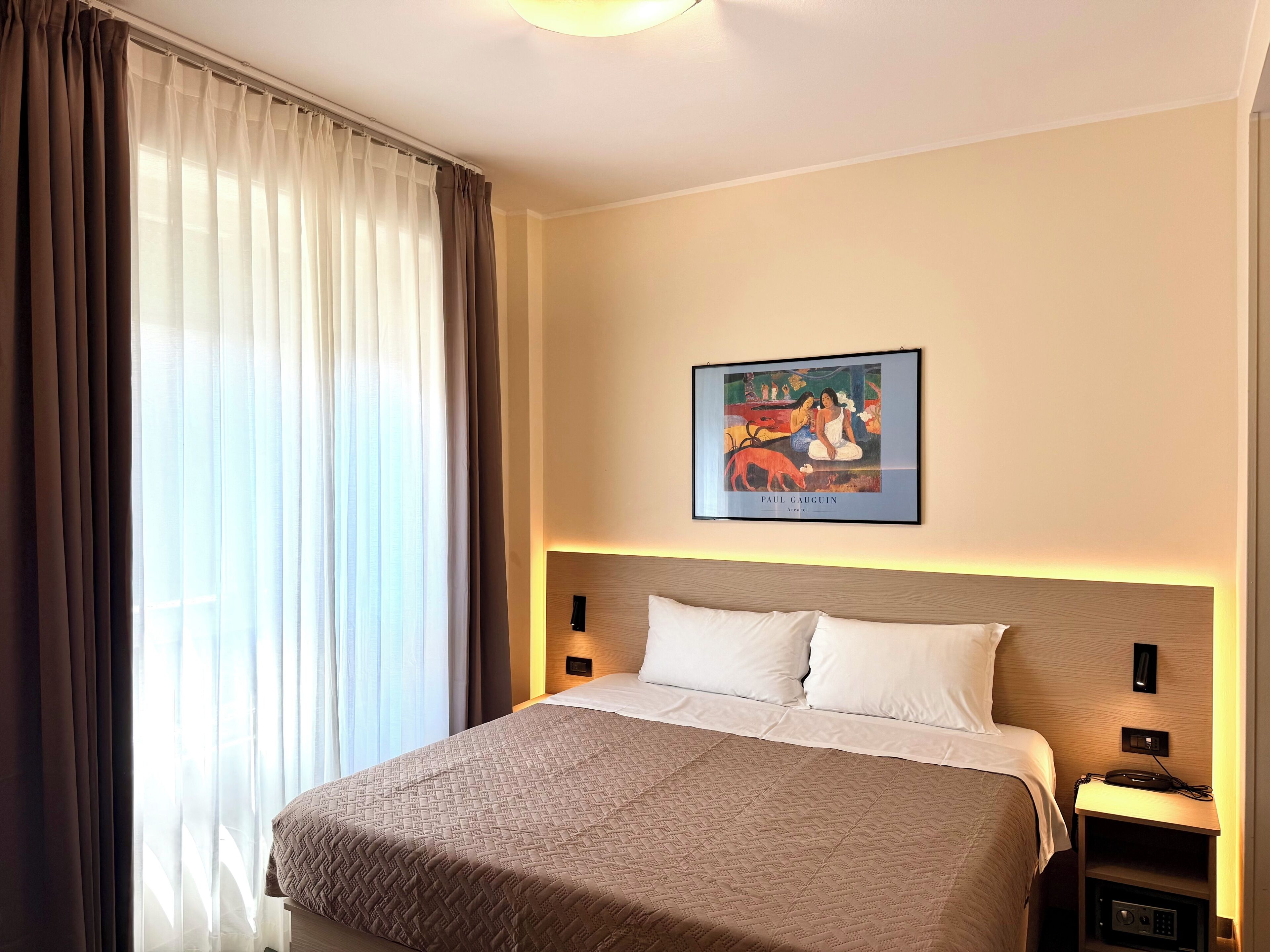 A hotel room with a bed, a nightstand, a wall-mounted painting, and a window with curtains.