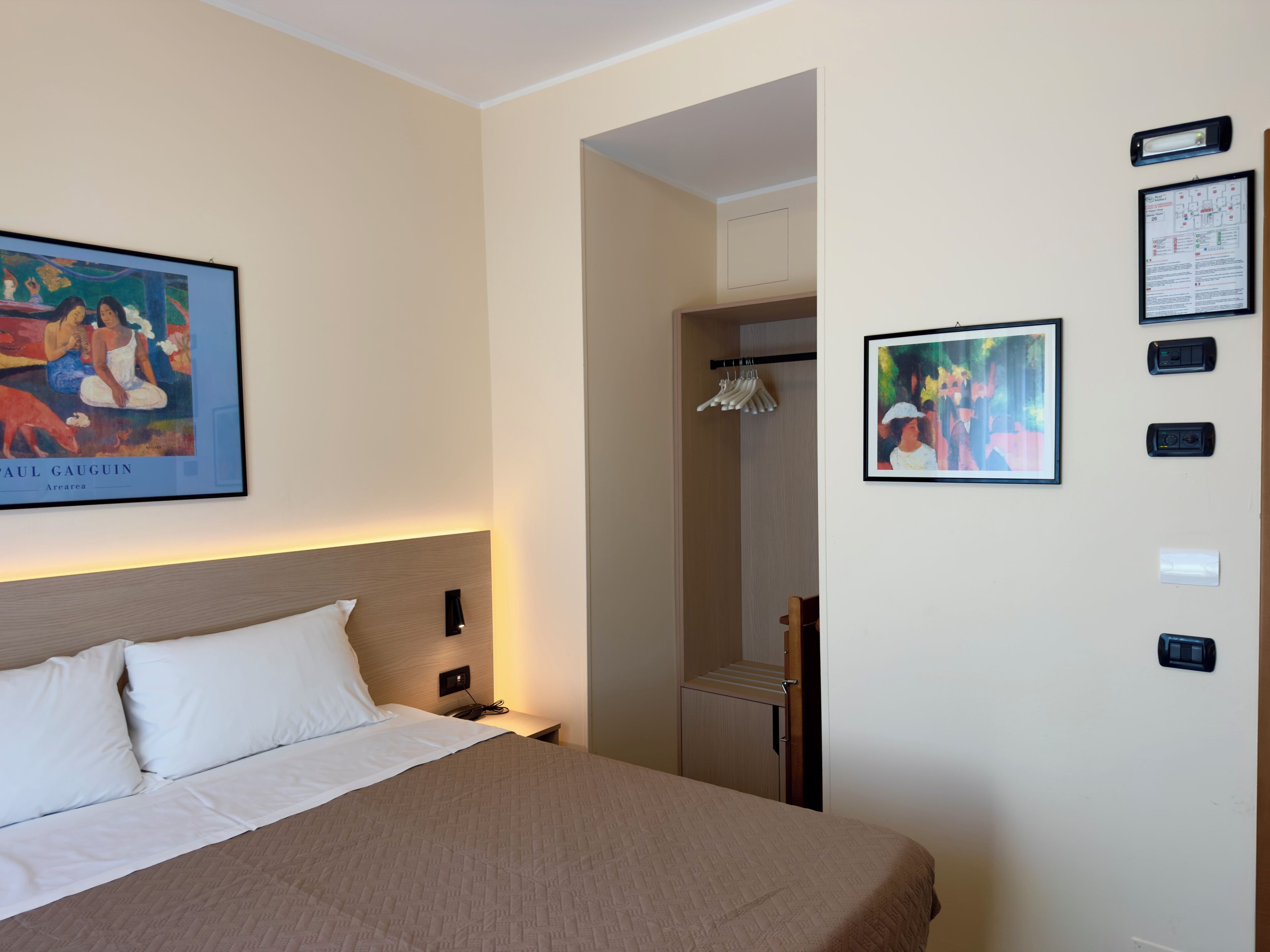 A hotel room with a bed, a painting, a wardrobe, and a wall-mounted information board.