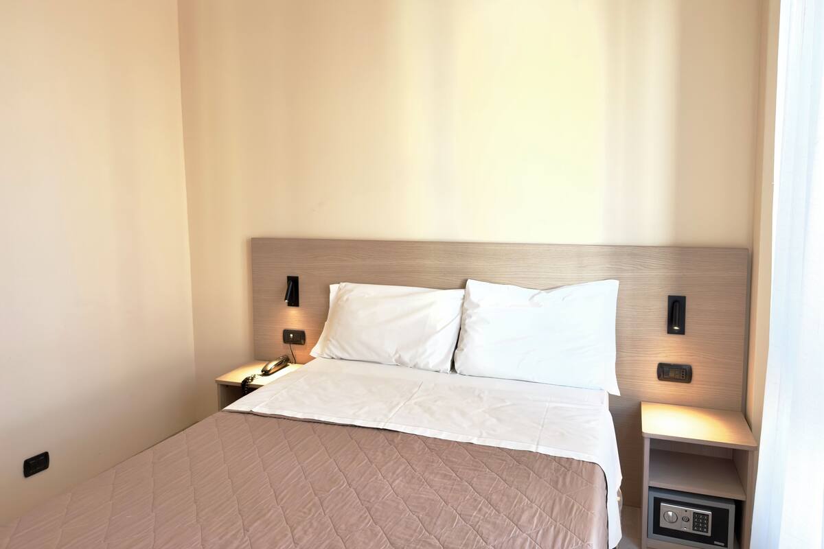 Ein ordentlich bezogenes Bett mit gesteppter Decke, ein Holzkopfstück, Nachttischlampen und ein Nachttisch mit Telefon und einem kleinen elektronischen Gerät.
