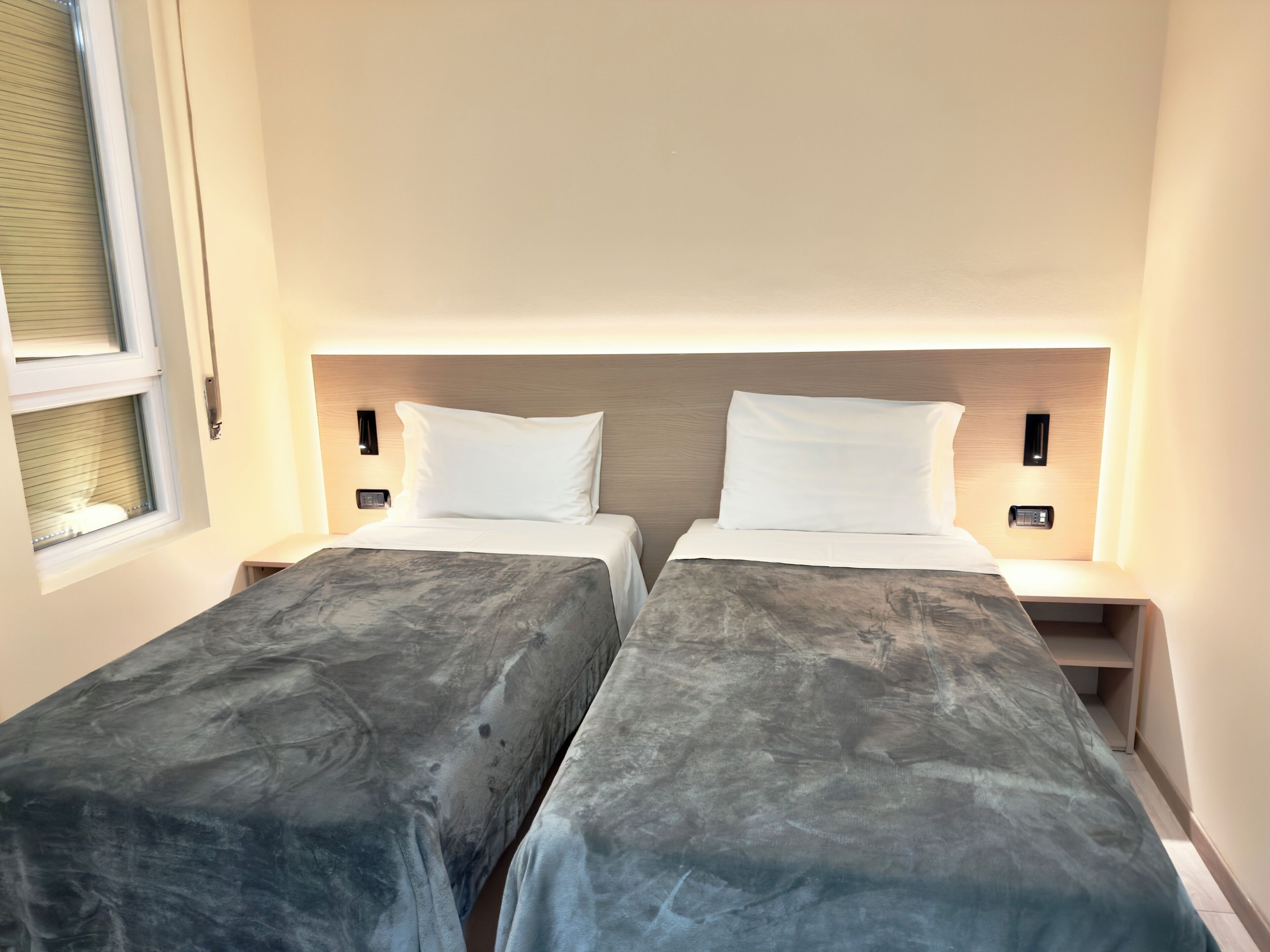 A hotel room with two single beds, each with a headboard, bedside tables, and a window with blinds.