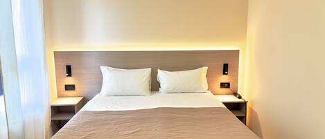 A hotel room with a large bed, two bedside tables, and a headboard with built-in lighting.