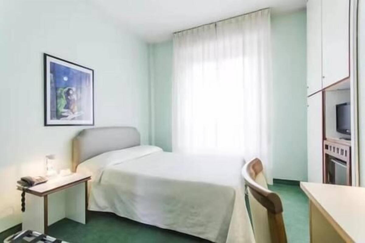 Ein Einzelbett mit weißer Bettdecke, ein Nachttisch mit Lampe, ein Fernseher auf einem Schrank und ein gerahmtes Bild an der Wand.