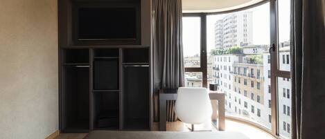 A hotel room with a large bed, a TV mounted on the wall, a desk, and a view of the city through large windows.
