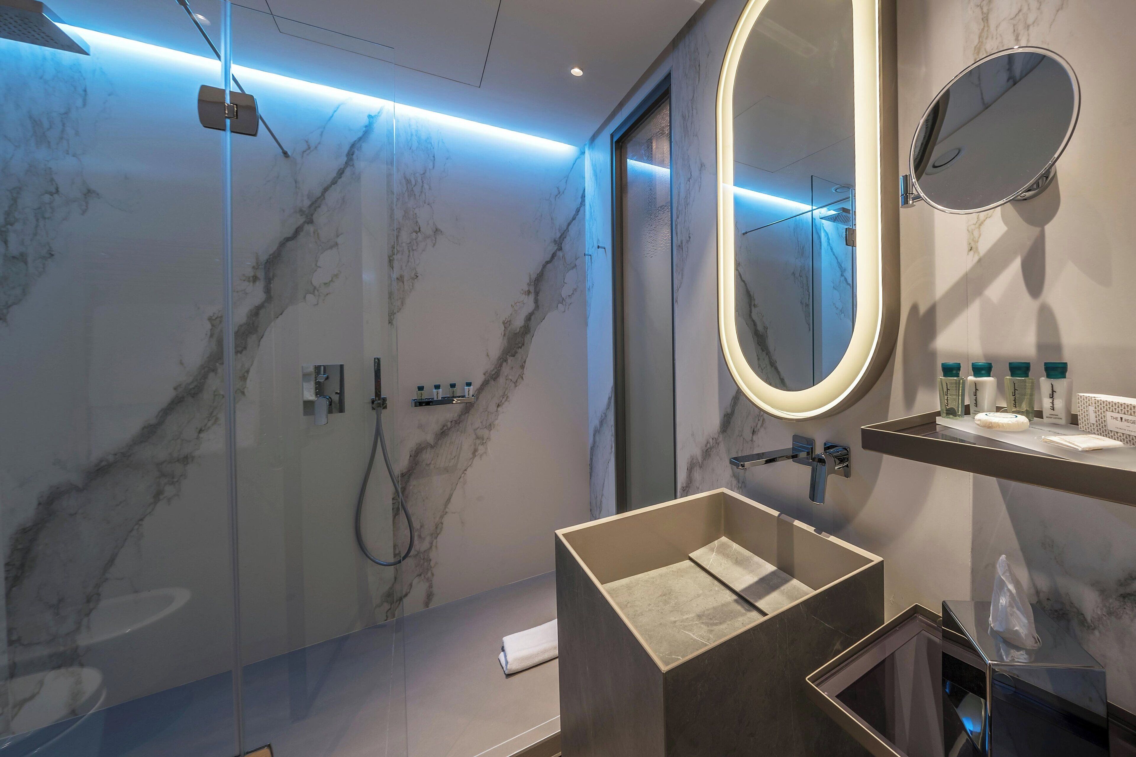 A modern bathroom with a glass-enclosed shower, a marble-tiled wall, a large mirror, and a vanity with toiletries.