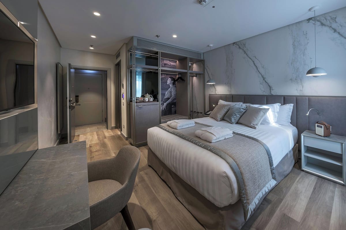 A modern hotel room with a large bed, a seating area, a wardrobe, and a television.
