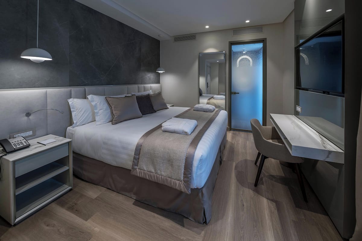 A modern hotel room with a large bed, bedside table with a phone, a desk with a chair, and a large mirror.