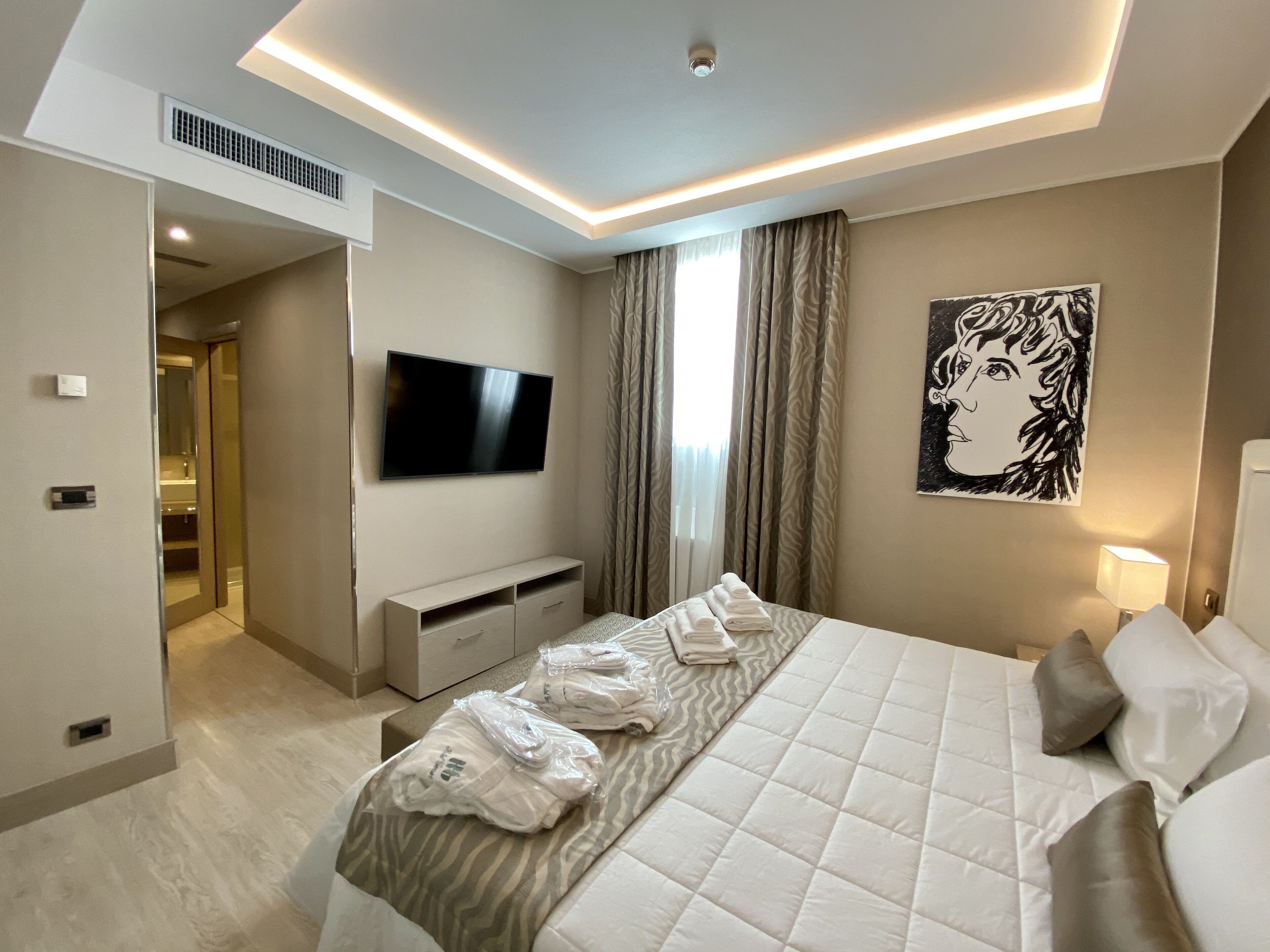 Une chambre d’hôtel moderne avec un grand lit, un téléviseur à écran plat fixé au mur, une table de chevet avec une lampe et un tableau au mur.