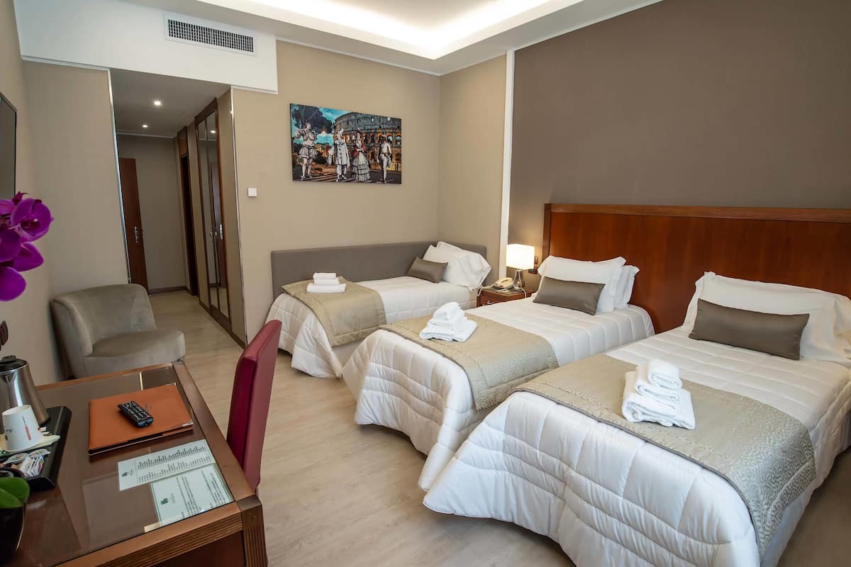 A hotel room with three beds, a desk with a laptop, a mirror, and a painting on the wall.