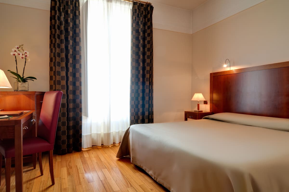 A hotel room with a bed, a desk with a chair, a lamp, and a window with curtains.