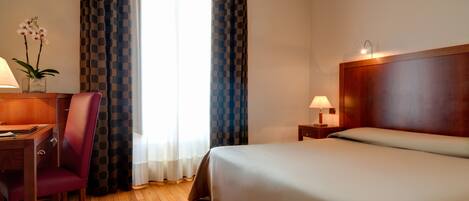 A hotel room with a bed, a desk with a chair, a lamp, and a window with curtains.