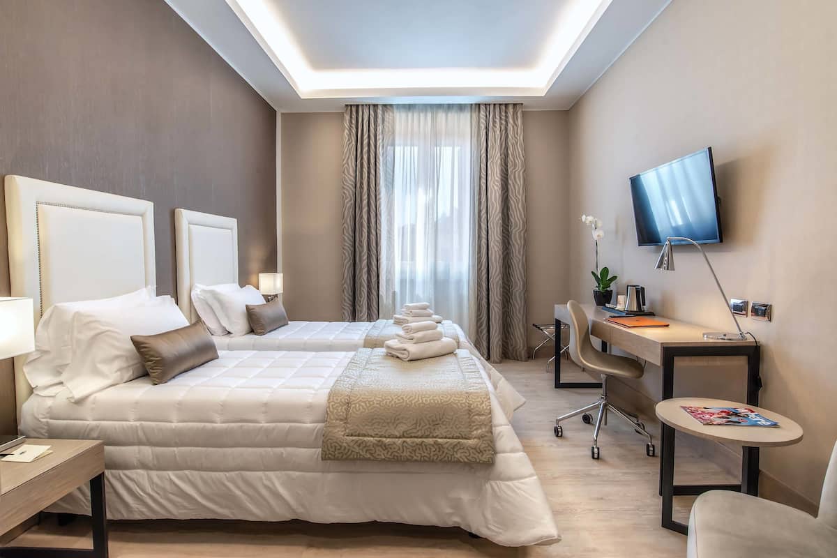 A hotel room with a large bed, a desk, a chair, a TV, and a window with curtains.