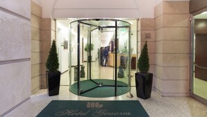 Une porte tournante avec un encadrement en verre, un paillasson vert sur lequel est inscrit le nom « Hôtel Genova », et deux plantes en pot.