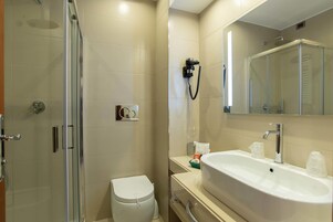 Et moderne badeværelse med et glasafskærmet brusebad, et hvidt toilet, en vask med spejl og en håndklædedispenser.