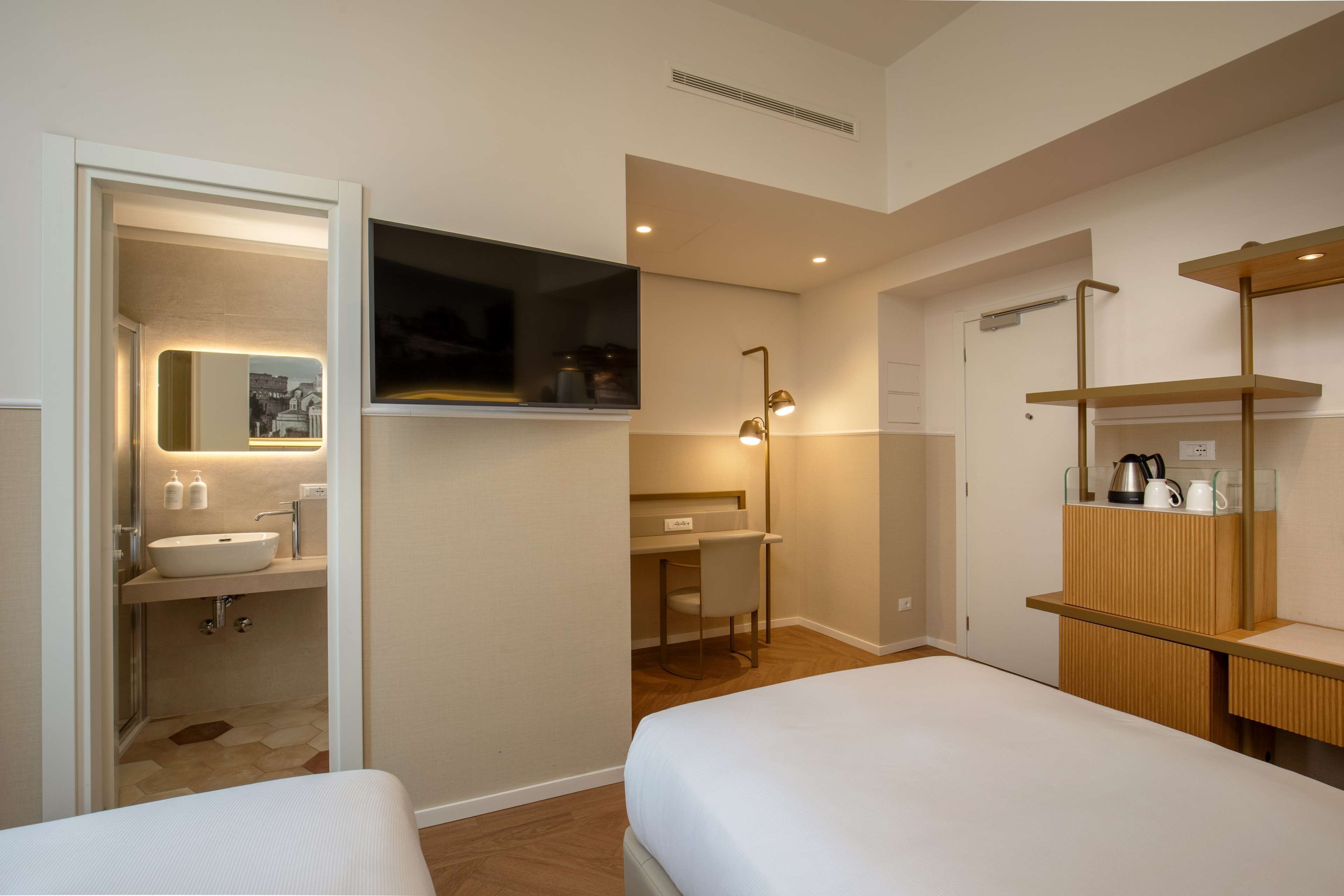 A modern hotel room with a large bed, a desk with a chair, a vanity with a mirror, and a shelf with toiletries.