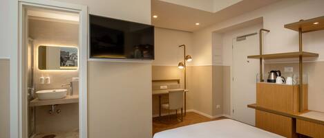 A modern hotel room with a large bed, a desk with a chair, a vanity with a mirror, and a shelf with toiletries.
