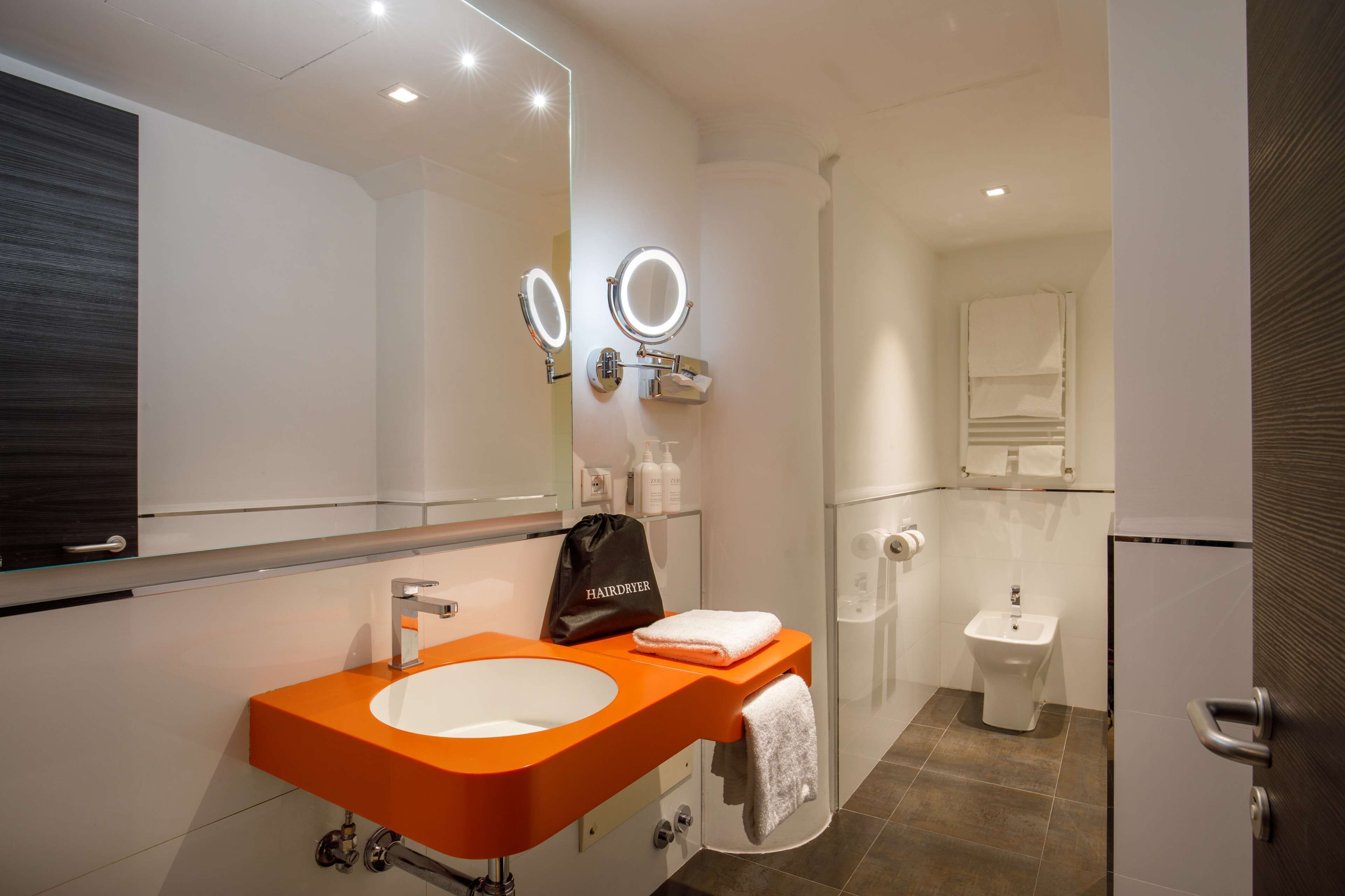A modern bathroom with a large mirror, two round mirrors, a sink with an orange countertop, a hairdryer bag, and a towel.