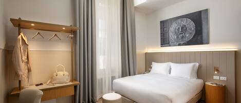 A modern hotel room with a bed, bedside table, chair, and a coat rack with hangers.