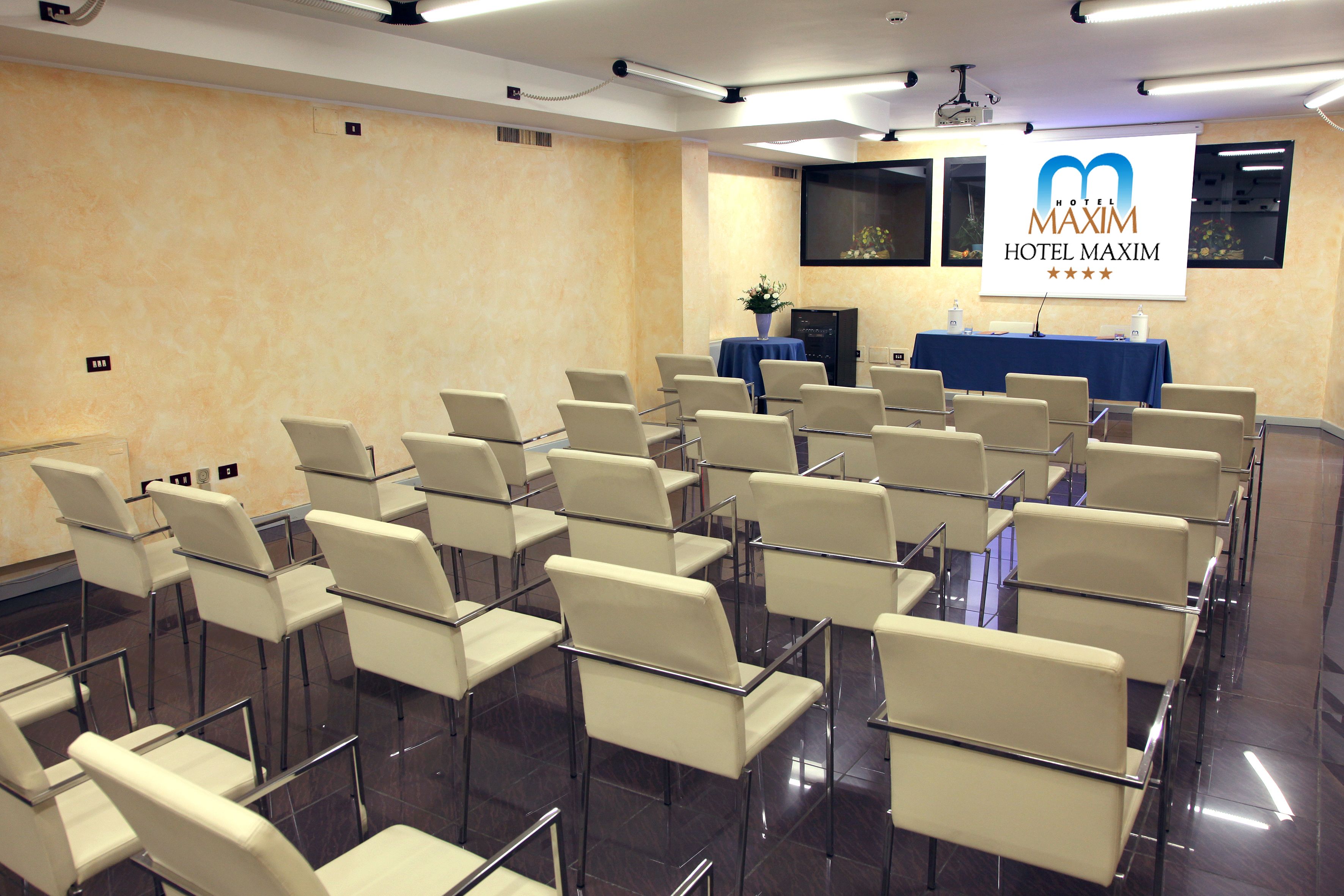 A conference room with rows of white chairs, a projector screen, and a presentation area with a table and chairs.