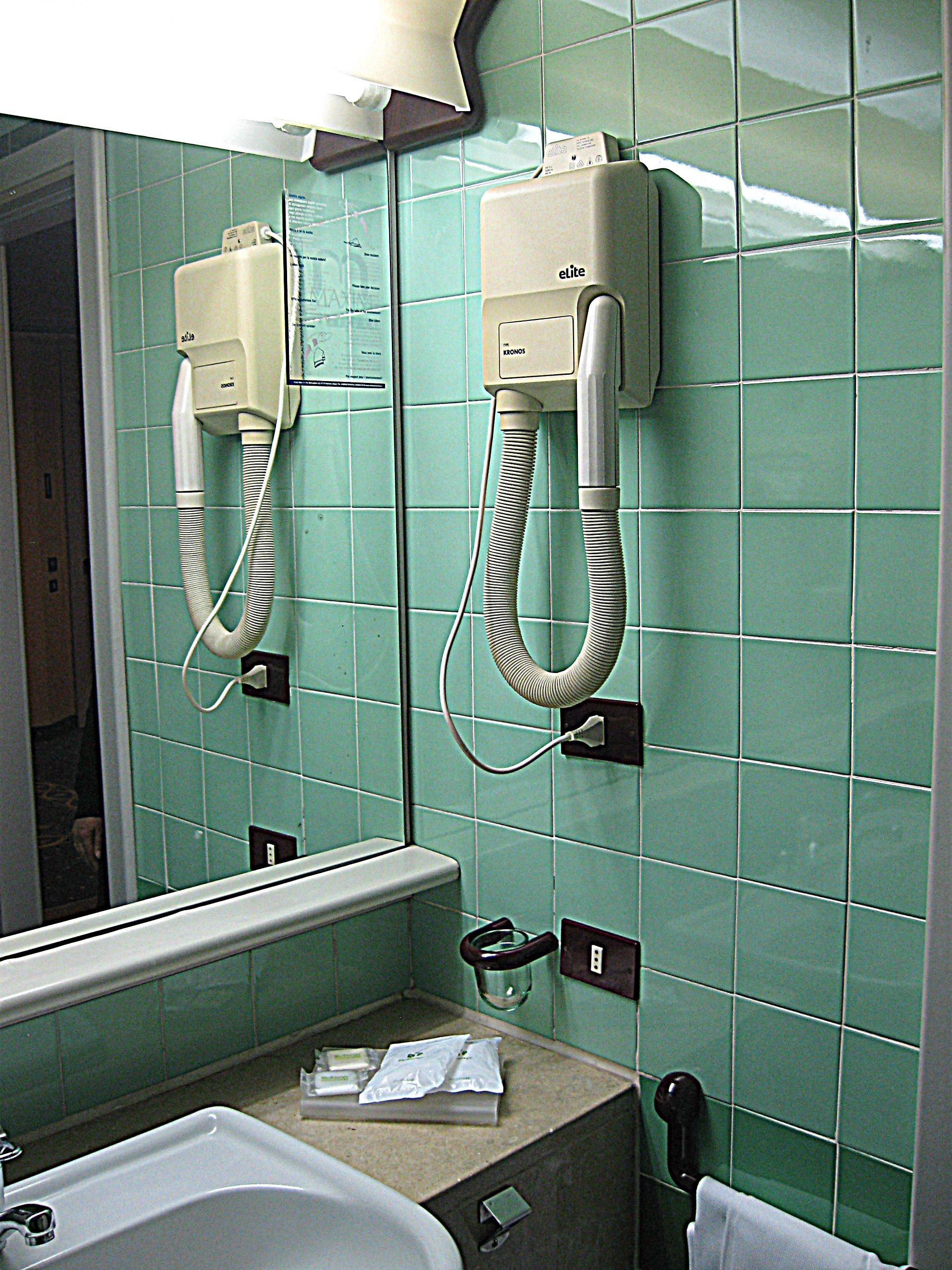 A bathroom with green tiles, a sink, and two hair dryers mounted on the wall.