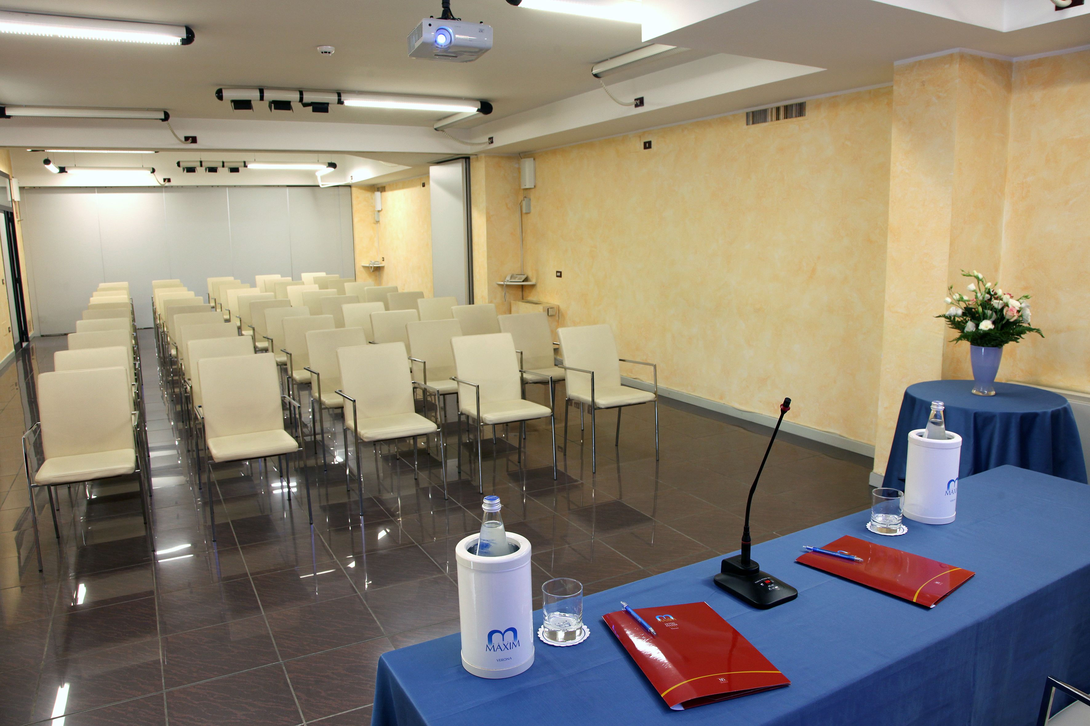 Ein Konferenzraum mit Reihen von Stühlen, einem Projektor, einem Tisch mit einem Mikrofon, einem Wasserspender und einer Vase mit Blumen.