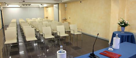 Uma sala de conferências com fileiras de cadeiras, um projetor, uma mesa com microfone, um bebedouro e um vaso com flores.