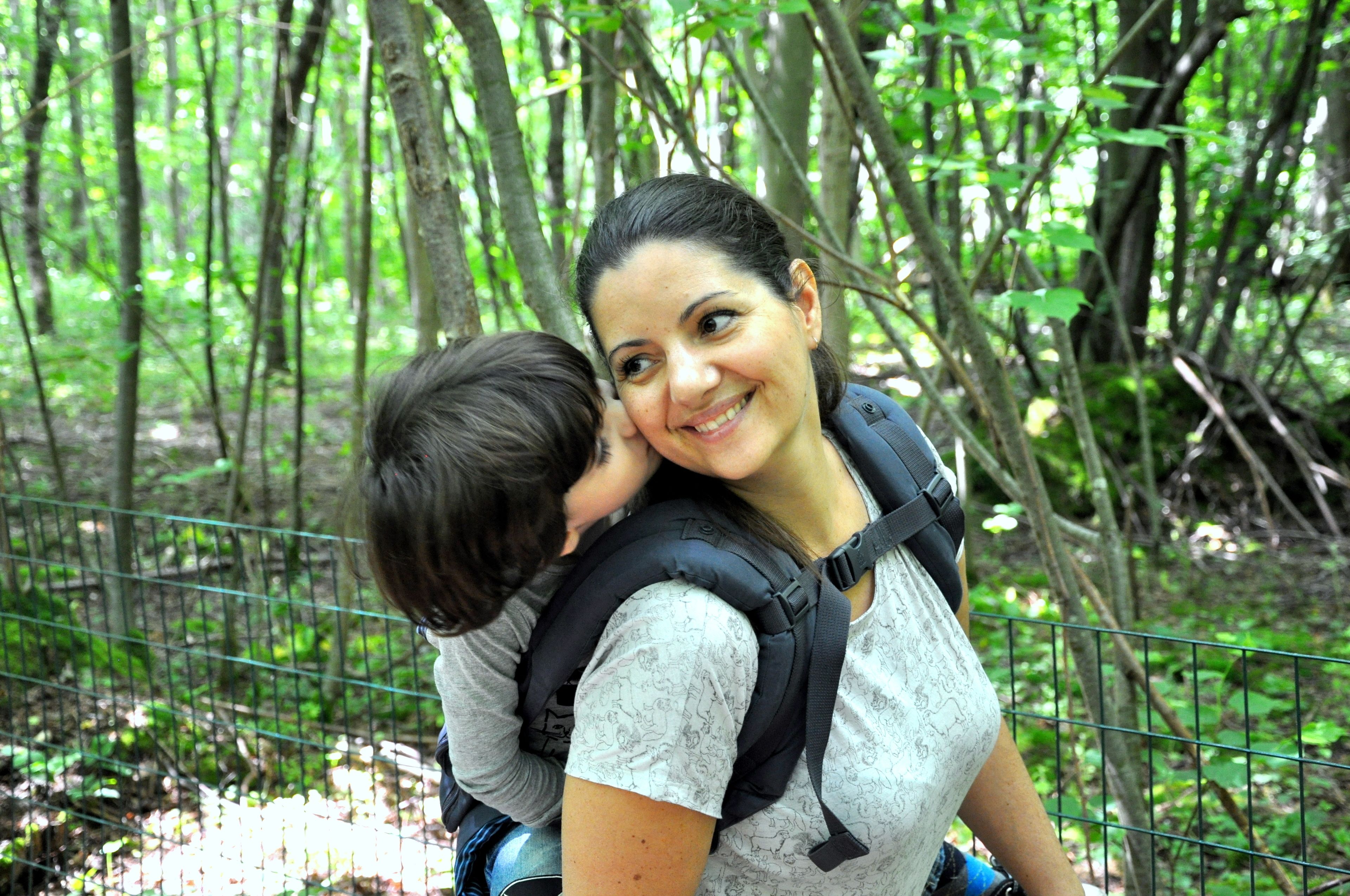 A person carrying a child on their back, surrounded by a forested area with a metal fence.