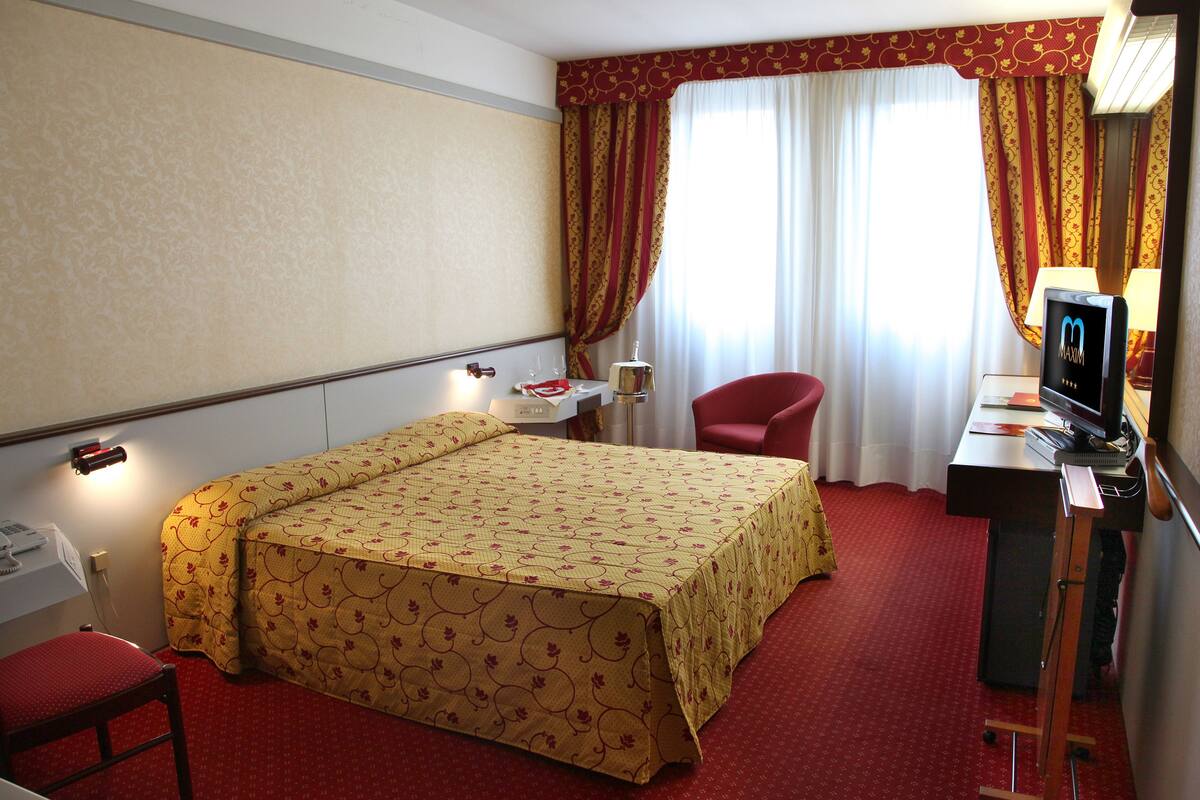 Ein Hotelzimmer mit einem Bett, einem Schreibtisch mit Computer, einem Sessel, einem Fernseher und einem Fenster mit Vorhängen.