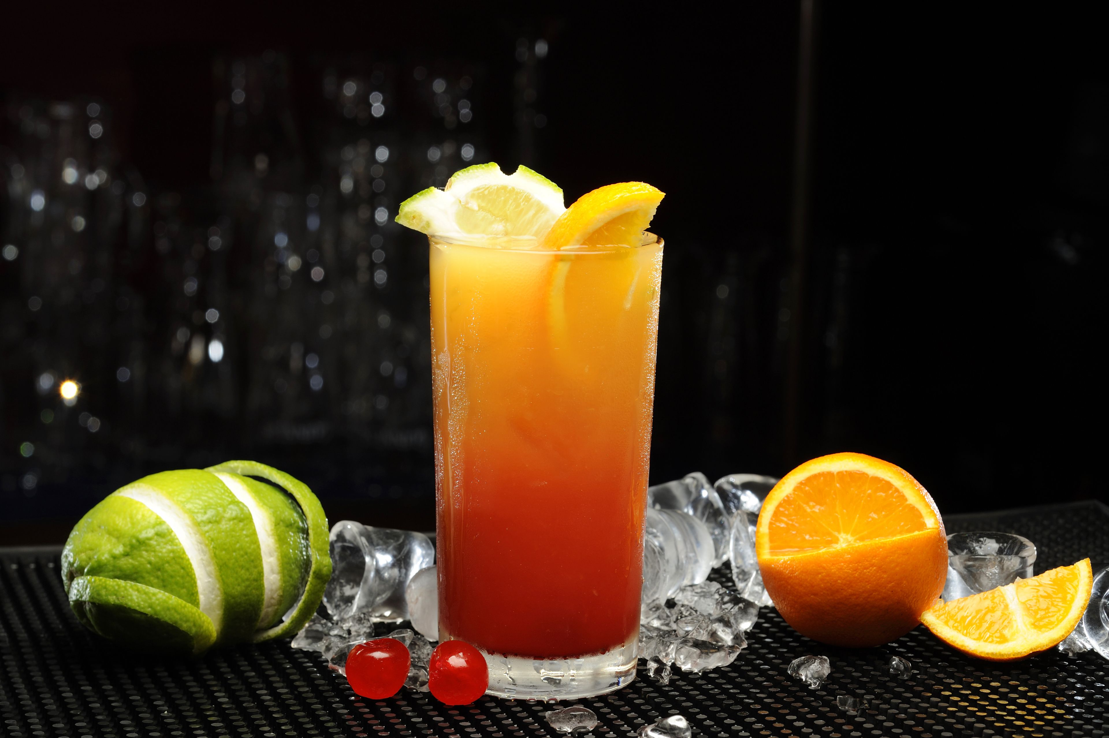 Un vaso alto con una bebida degradada, una lima, una naranja y cubitos de hielo sobre una superficie texturizada.