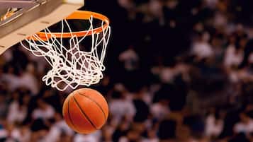 Una canasta de baloncesto con red, un balón de baloncesto en pleno vuelo y un público desenfocado al fondo.
