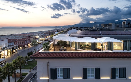 A coastal hotel with a terrace overlooking the beach, a dining area with umbrellas, and a view of the sea and mountains.