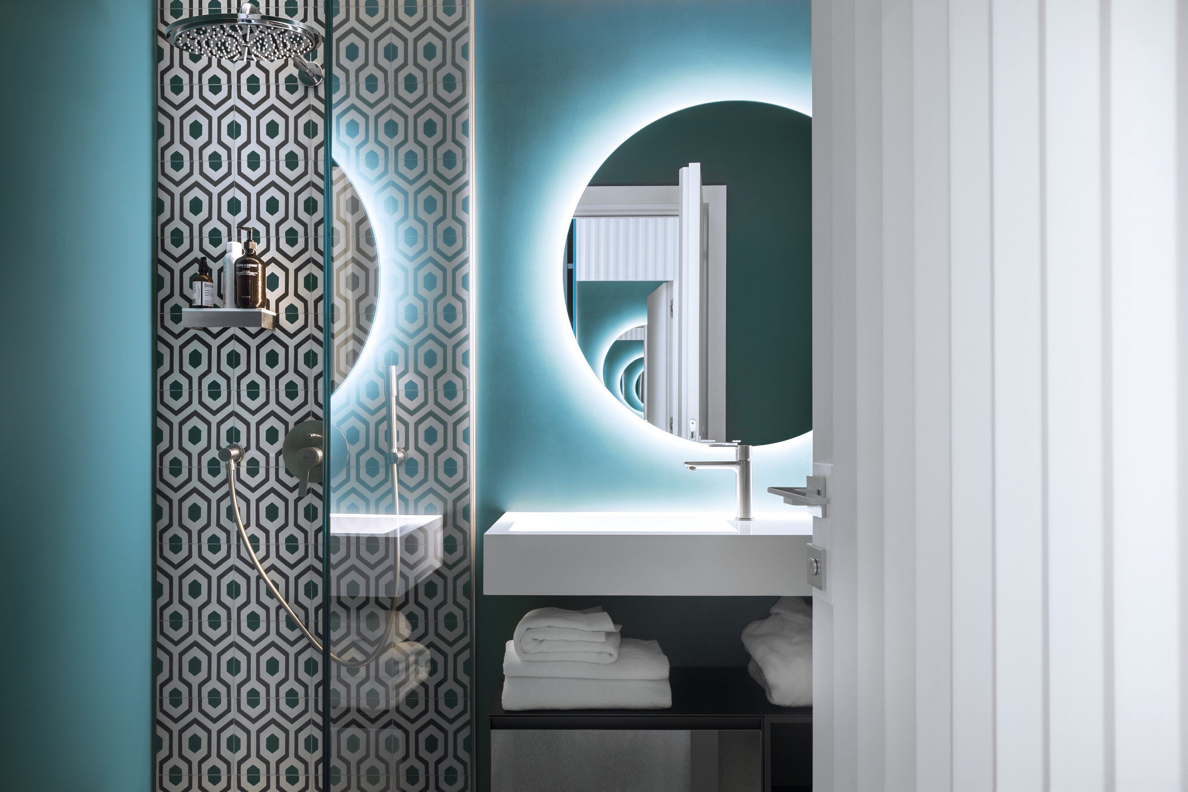 A modern bathroom with a geometric patterned wall, a round mirror, a white sink, and neatly folded towels.