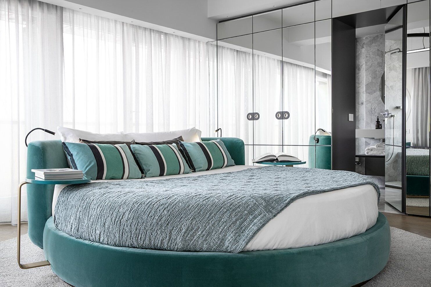 A modern bedroom with a teal bed, striped pillows, a bedside table with a book, and a large mirror.